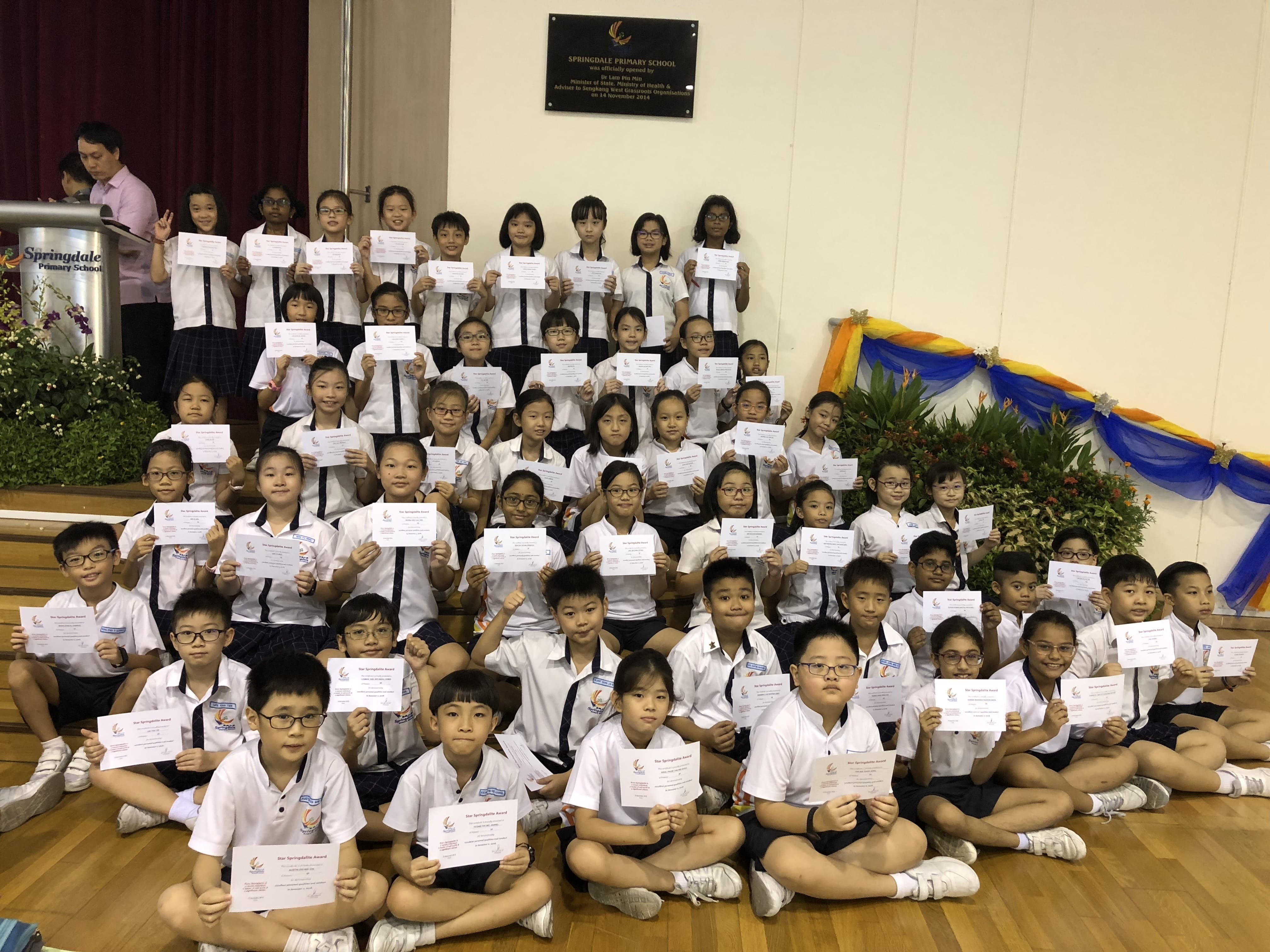 Large group of Springdale Primary School students in uniform holding award certificates on a school hall stage.